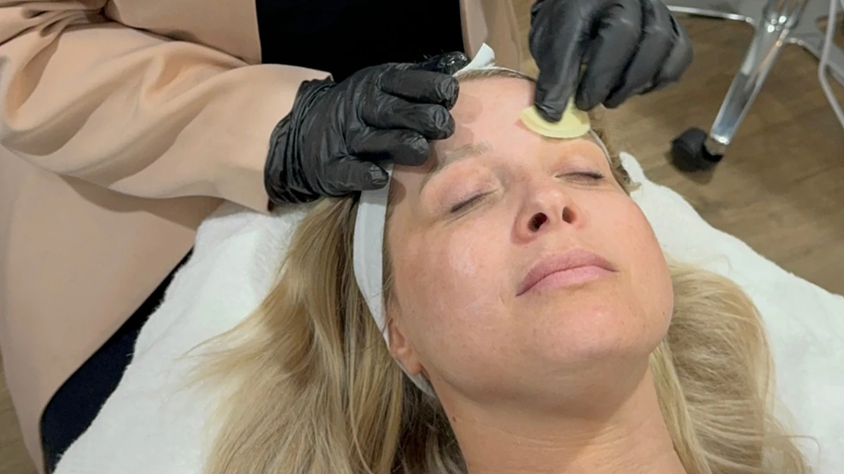 A woman with blonde hair and eyes closed is lying on a white towel. An esthetician wearing black gloves applies a facial treatment with a sponge, conveying a calm and soothing atmosphere.