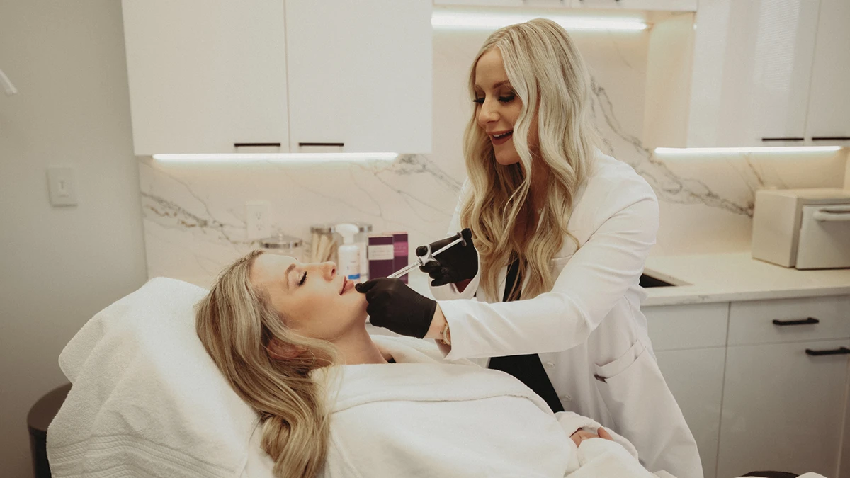 A woman in a white coat and gloves performs a cosmetic procedure on a relaxed woman lying on a treatment bed in a well-lit, modern clinic.