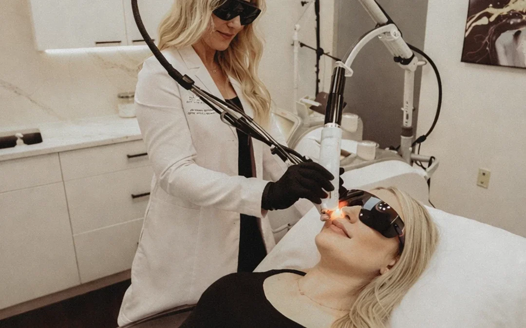 A woman in a white coat and black gloves uses a laser device on a patient's face. Both wear protective eyewear. The setting is a clinical treatment room.