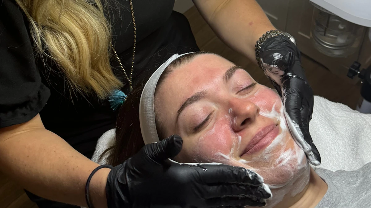 Woman receiving a facial, eyes closed and smiling, as skincare professional with blonde hair applies cream with gloved hands, conveying relaxation.