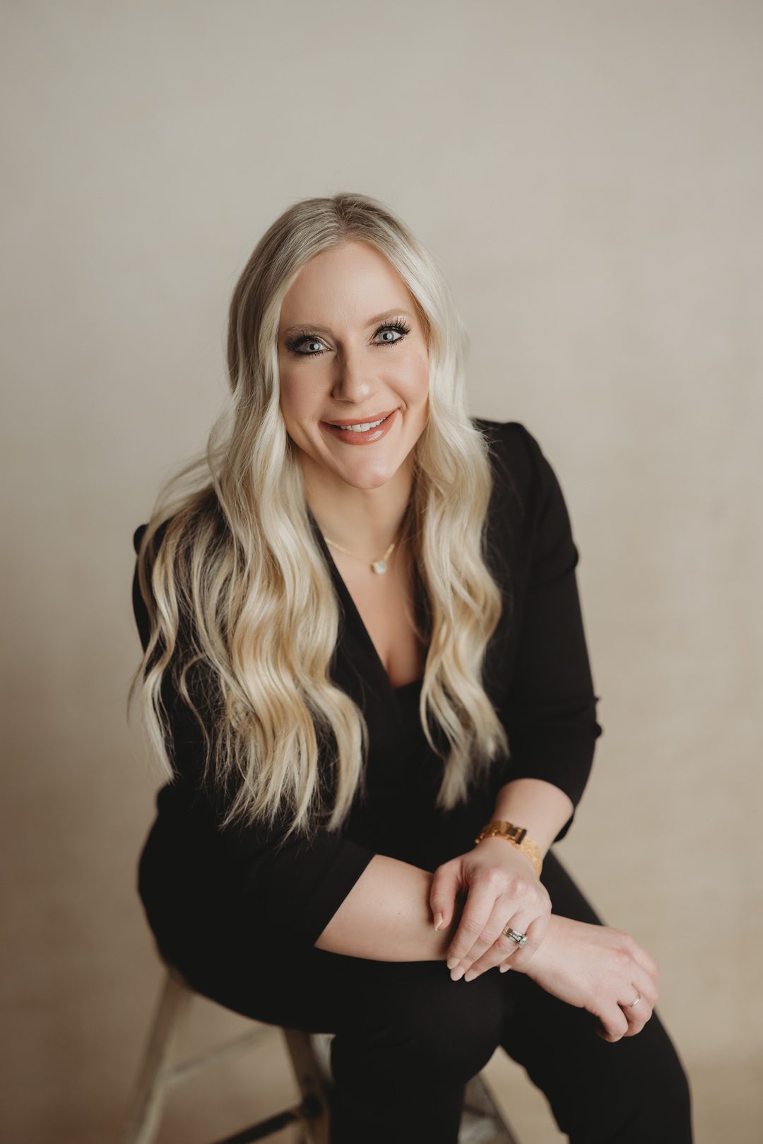 Blonde woman with wavy hair and striking eyes wearing a black sleeveless top, looking confidently over her shoulder against a plain white background.
