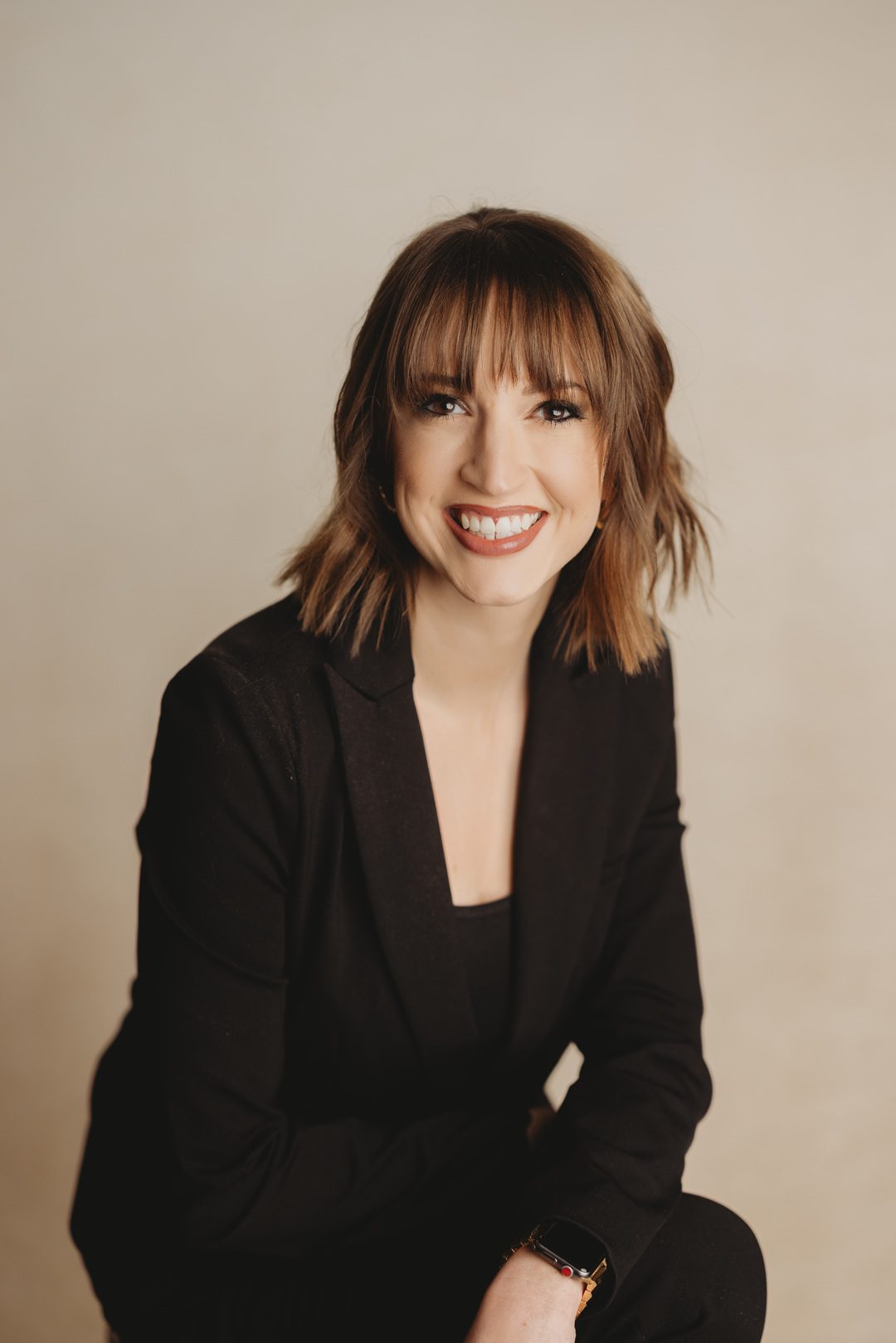 Smiling woman with long brown hair and bangs, wearing a navy blazer, against a neutral background. The tone is professional and welcoming.