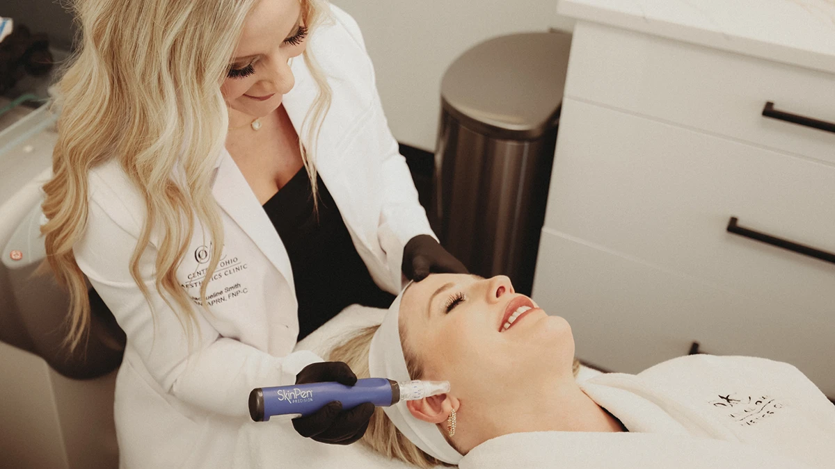 A smiling aesthetician with long blonde hair applies a Skincare treatment device to a relaxed client's face. The setting is clean and professional.