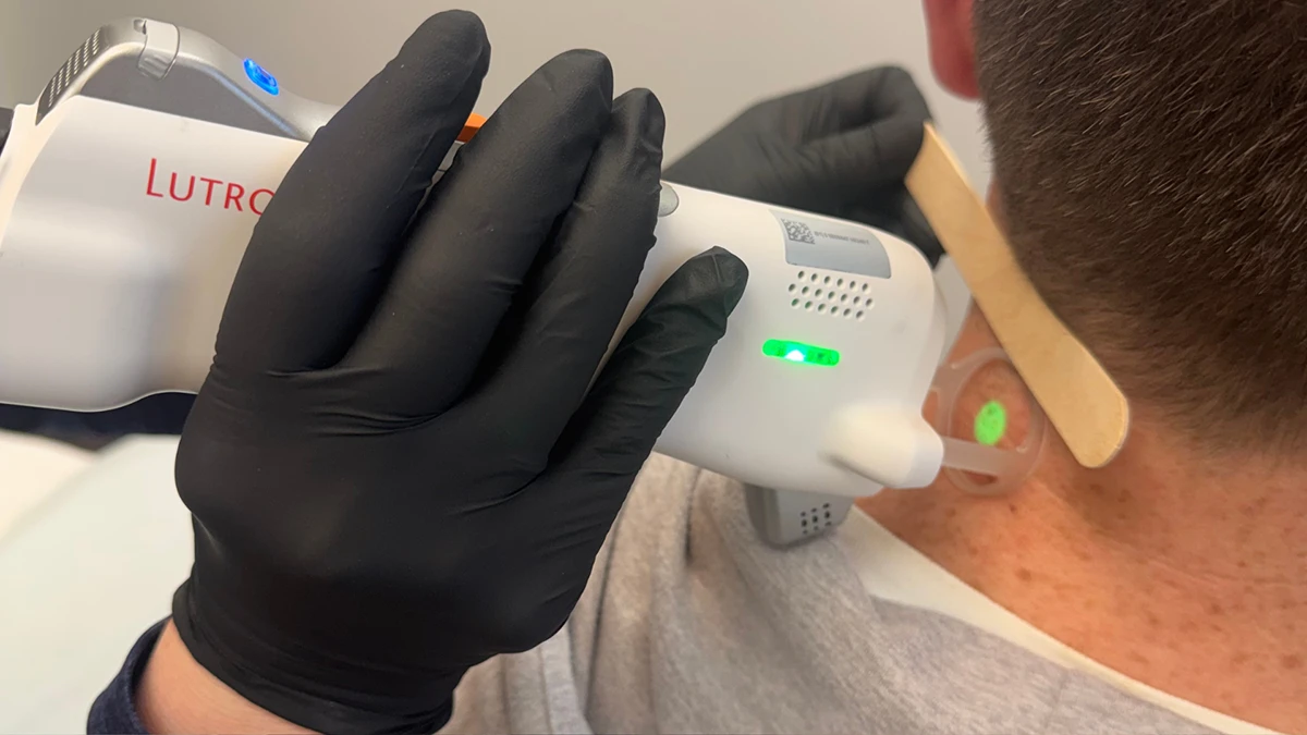 A gloved hand holds a laser device near a person's neck, using a tongue depressor for guidance. The setting appears clinical and focused. A gloved hand holds a laser device near a person's neck, using a tongue depressor for guidance. The setting appears clinical and focused.