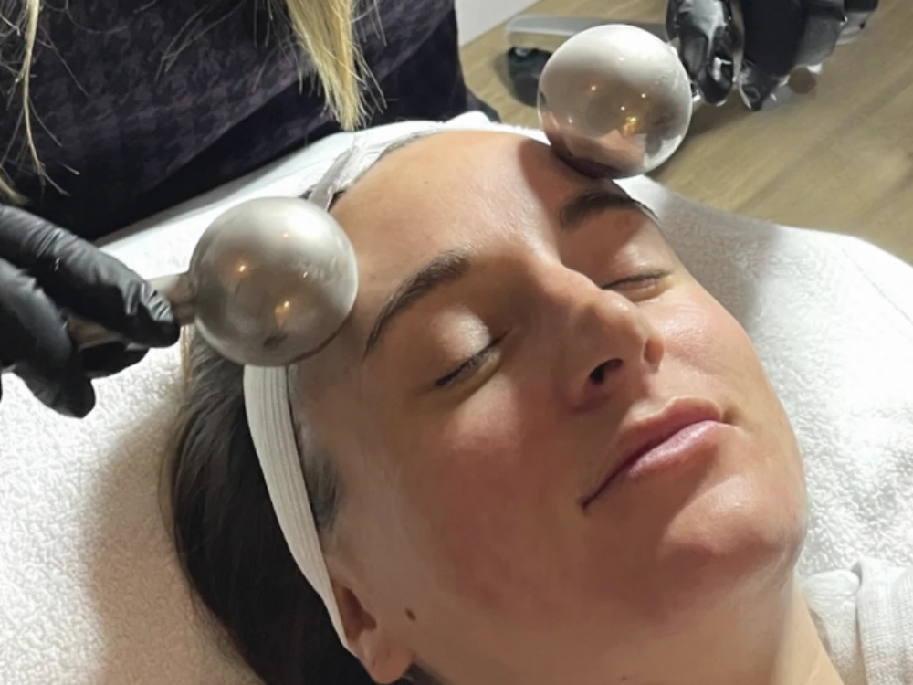 Woman receiving a relaxing facial treatment with two metal globes held by gloved hands on her forehead. Her eyes are closed, and she is serene.