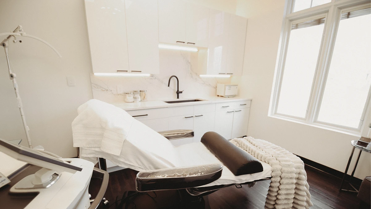 Modern, bright spa room with a reclining chair draped in a white blanket, a sleek black faucet and sink, and large windows letting in natural light. Modern, bright spa room with a reclining chair draped in a white blanket, a sleek black faucet and sink, and large windows letting in natural light.