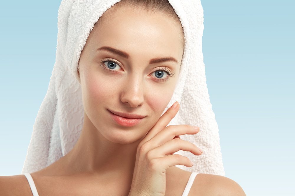 Close up shot of young Caucasian female with blue eyes and fresh healthy skin with no makeup, looking at the camera with beautiful smile while getting facial treatment in spa salon. Beauty concept